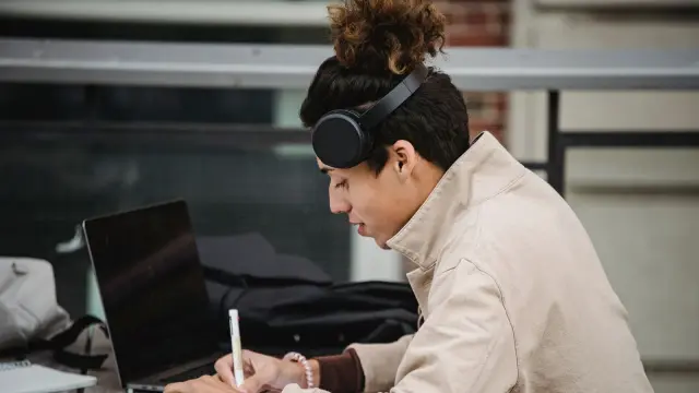 Estudiante con auriculares de diadema trabajando con el portátil en una mesa de biblioteca